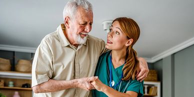 A nurse supports an elderly man standing beside a wheelchair in a cozy living room.