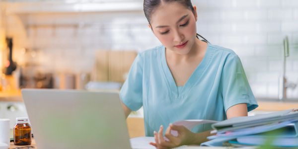 A nurse studies documents while sitting at a desk with a laptop and medical supplies.