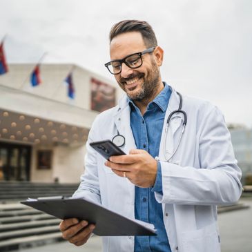 Smiling male doctor using phone and clipboard outdoors.