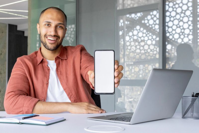 Smiling man holding smartphone displaying screen at desk with laptop and notebook. Shows use of technology and business in modern office environment. Ideal for communication concept.