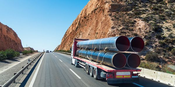 A truck carrying large metal pipes on a highway through rocky terrain.