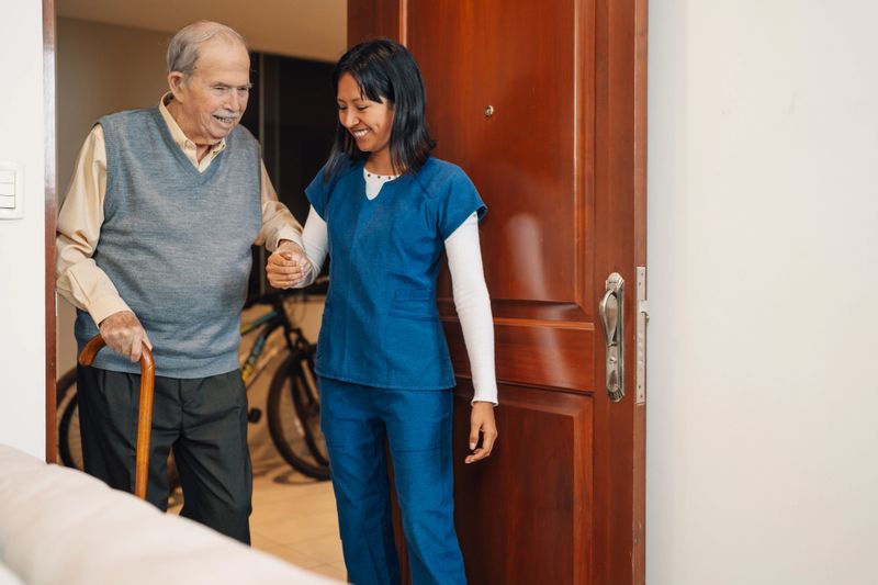 Old man assisted by a nurse that helps him to walk during a home visit
