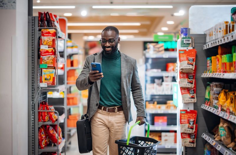 A cheerful man carrying a basket shops in a grocery store while browsing on his smartphone, demonstrating a modern lifestyle and technology use in everyday activities.