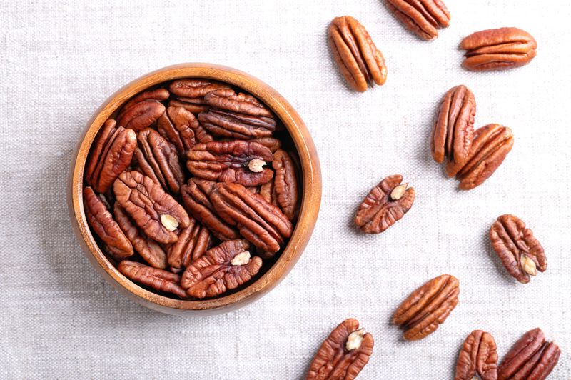 Pecan halves in a wooden bowl on linen fabric. Shelled and dried pecan nuts, seeds and fruits of Carya illinoinensis. Ready to eat fresh or roasted as snack, and used for baking. Close-up from above.