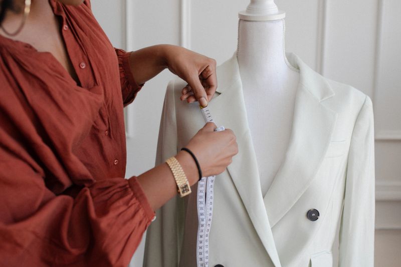 A black woman fashion designer measuring a white blazer on a dress form. This fashion designer focuses on precision in tailoring, showcasing her skills in custom clothing design.