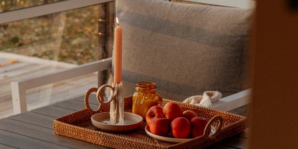 Wicker tray with lit candle, jar, and peaches on a cozy chair by the window.