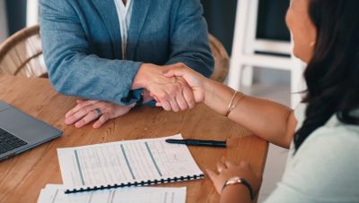 Two people shaking hands over a desk with documents and a laptop.
