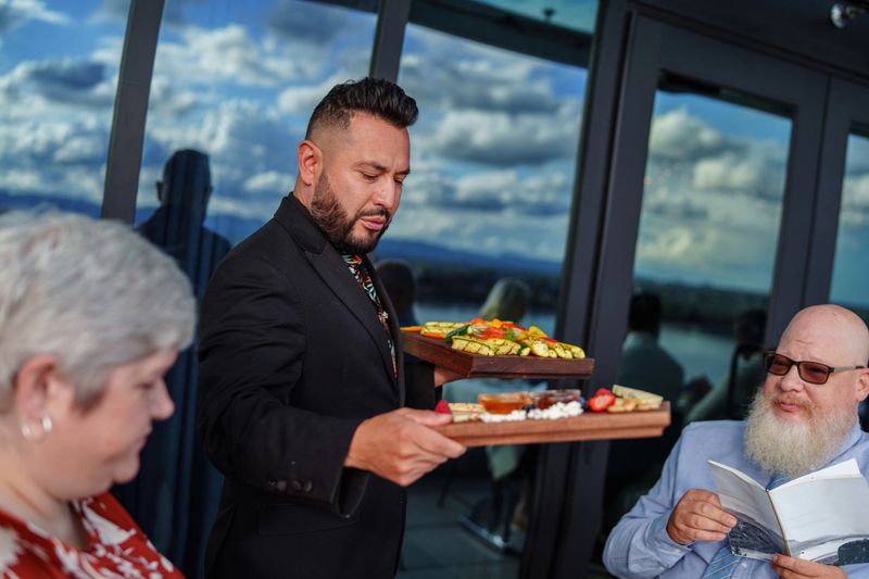 A male waiter of Hispanic descent serves charcuterie board appetizer platters to senior adult guests seated on the rooftop terrace of an upscale restaurant.