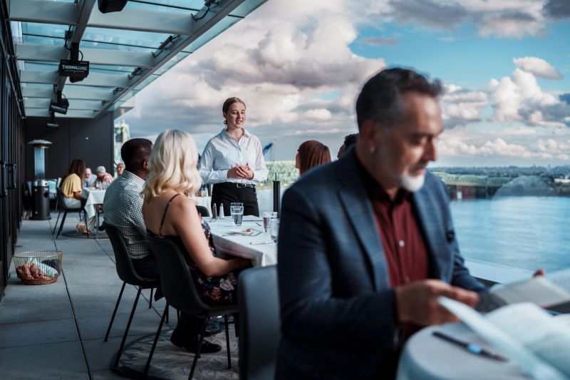 A restaurant hostess chats with a multiracial group of friends dining together on the rooftop terrace of an upscale restaurant on a cloudy yet warm evening.