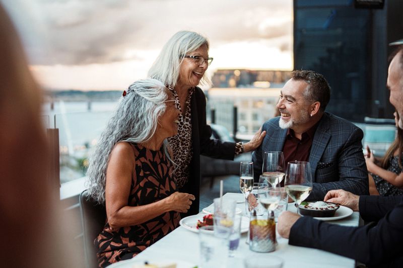 An active and beautiful senior woman smiles while saying goodbye to her friends after dining out together on the rooftop patio of an upscale restaurant with scenic river views at sunset.
