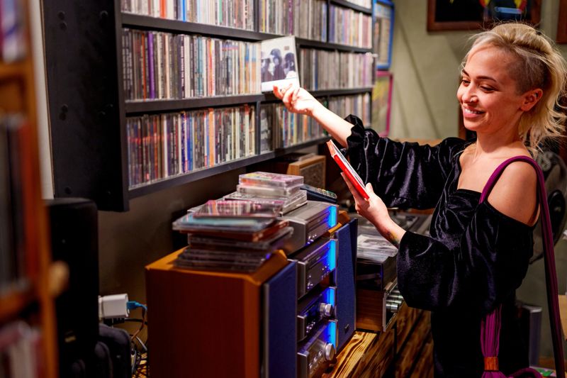 Young woman browsing CDs in a retro-style record store. Dressed in a chic off-the-shoulder black dress, she stands next to wooden shelves full of records, looking intently at one record. The interior of the shop has a vintage atmosphere, decorated with framed posters, CDs and colorful memorabilia, giving off a nostalgic vibe. Perfect for themes of music, nostalgia, vintage shopping and record culture.