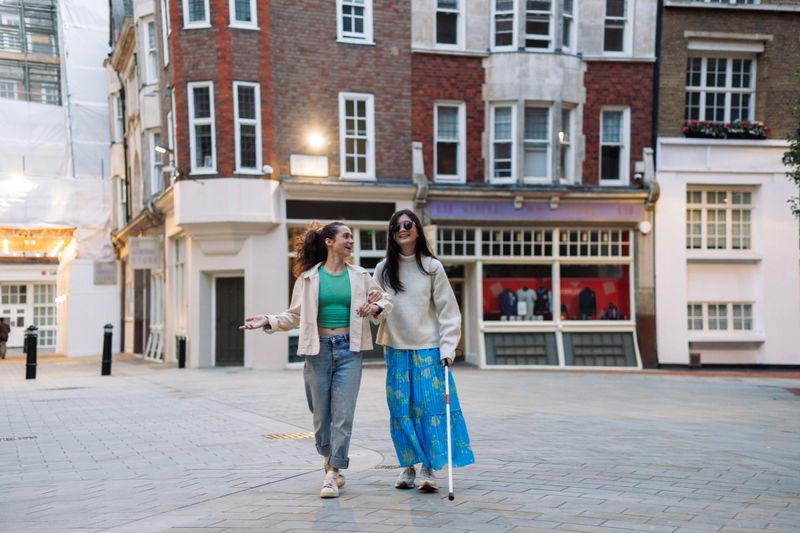 Two happy female friends walking together in the city, one of them helping her blind friend