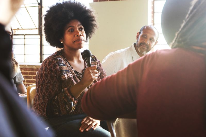 Black woman holding a microphone speaks in a group setting, with others listening attentively in a well-lit room with large windows. Black woman talking in microphone at a support and community event.