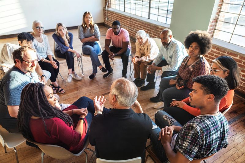Diverse group in a circle, discussing in a bright room. Mixed genders, engaged in conversation, sharing ideas, and listening attentively. Diverse people sitting in a circle of support from community.