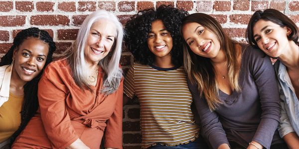 Five diverse women smiling together against a brick wall.