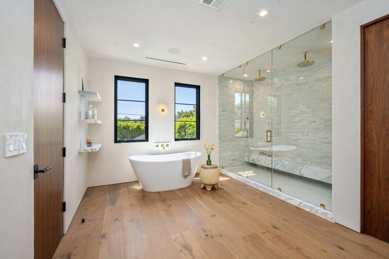 Modern bathroom with freestanding tub, glass shower, and wooden floor, featuring natural light from large windows.
