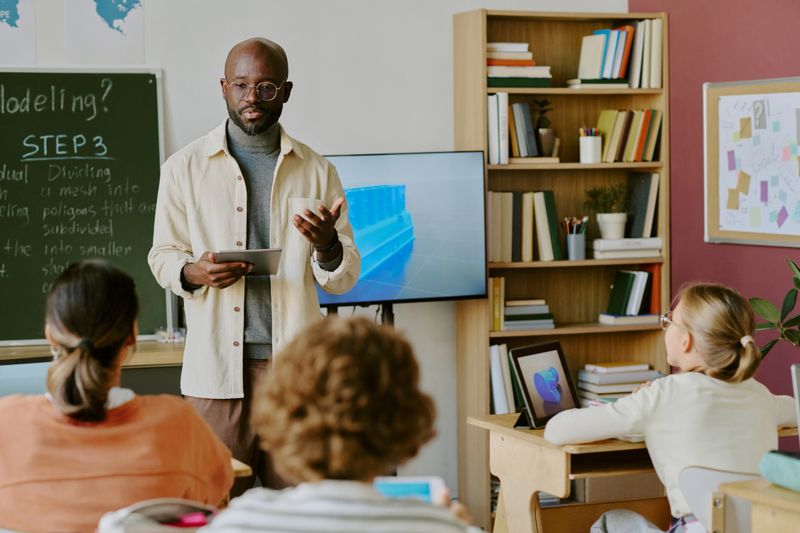 Experienced teacher using tablet leading group discussion in classroom with students attentively listening while presenting on flat screen TV