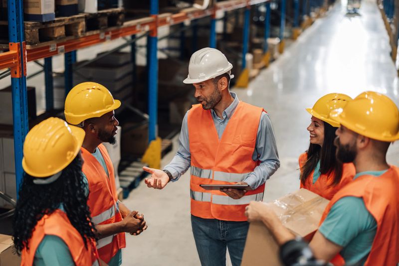 Warehouse manager holding a tablet is explaining logistics to a diverse group of workers wearing safety vests and hard hats