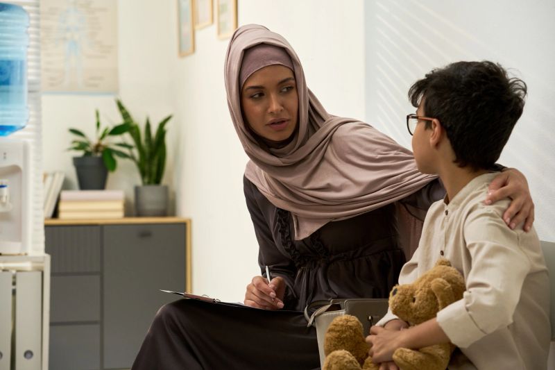 Muslim medical professional sitting with child in clinic, wearing hijab and holding clipboard. Engaging discussion hints at comforting medical consultation for young boy with teddy bear