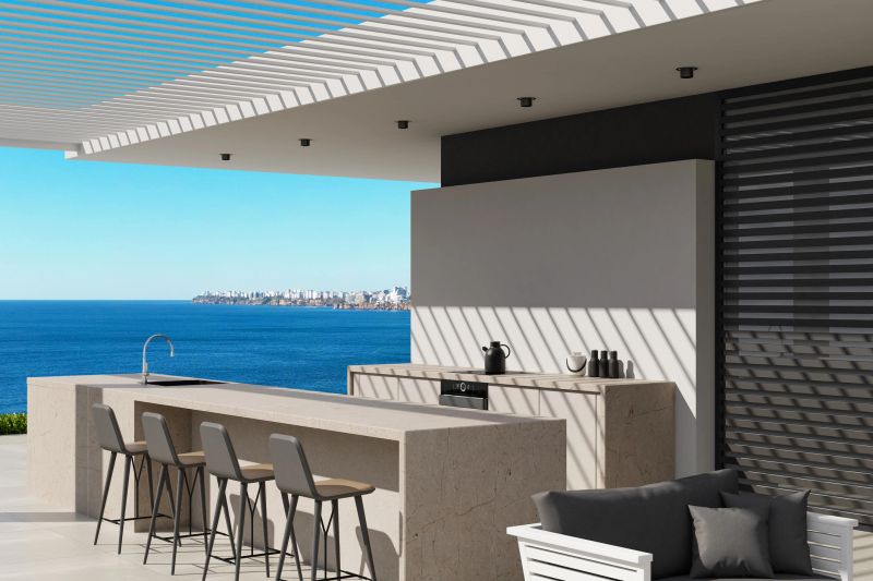 Immerse yourself in the charm of coastal elegance with this stunning image of a chic outdoor kitchen on a spacious patio terrace in Antalya, Turkey. The area features large white stone tiles and a stylish metal roof that provides shade, creating an inviting space for gatherings. The stone outdoor kitchen boasts a generous island counter with stools, perfect for socializing and culinary delights. Cozy white furniture enhances the atmosphere, inviting relaxation and comfort. With breathtaking views of the sea in the background and a harmonious palette of white and blue, this eco-friendly yet luxurious setting is ideal for promoting upscale outdoor living designs or inspiring architectural projects.