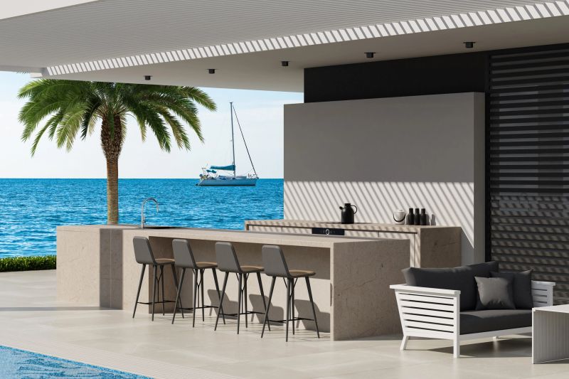 Discover the allure of coastal living with this captivating image of an elegant outdoor kitchen situated on a spacious patio terrace in Antalya, Turkey. The design features large white stone tiles and a sleek metal roof that offers ample shade, creating a perfect blend of style and functionality. The outdoor kitchen showcases a generous island counter with stools, ideal for entertaining, while the inviting white furniture adds a cozy touch. A shimmering swimming pool lies adjacent to the patio, enhancing the luxurious atmosphere. With panoramic views of the serene sea and a palette of white and blue, this eco-friendly retreat is perfect for showcasing luxury lifestyle projects or inspiring outdoor living spaces.