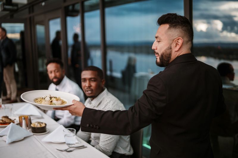 A well-dressed Latino waiter serves entrees to a table of male guests who are seated outside on the rooftop terrace of an upscale restaurant.