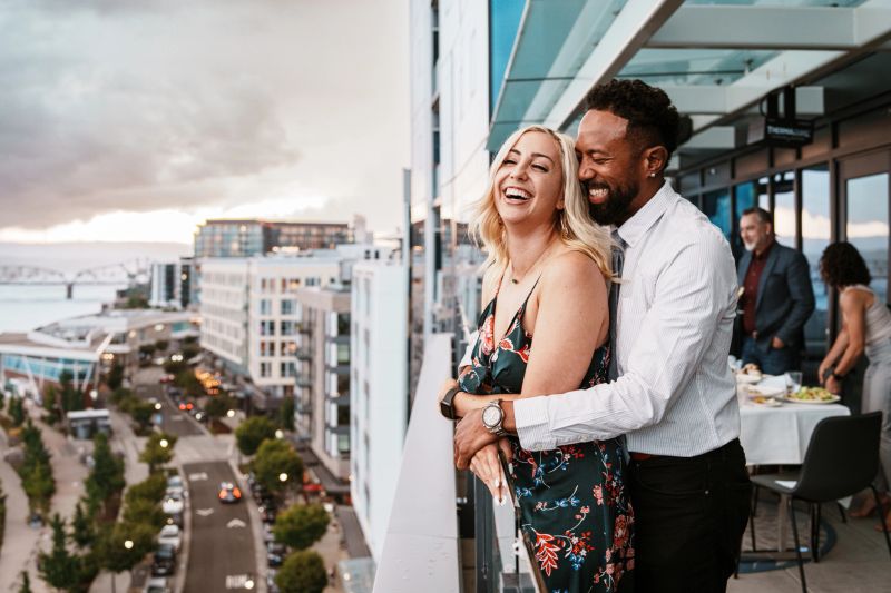 Profile view of a Black man and his Caucasian girlfriend affectionately embracing and enjoying scenic sunset views of the city from a restaurant's rooftop terrace while on a romantic date.