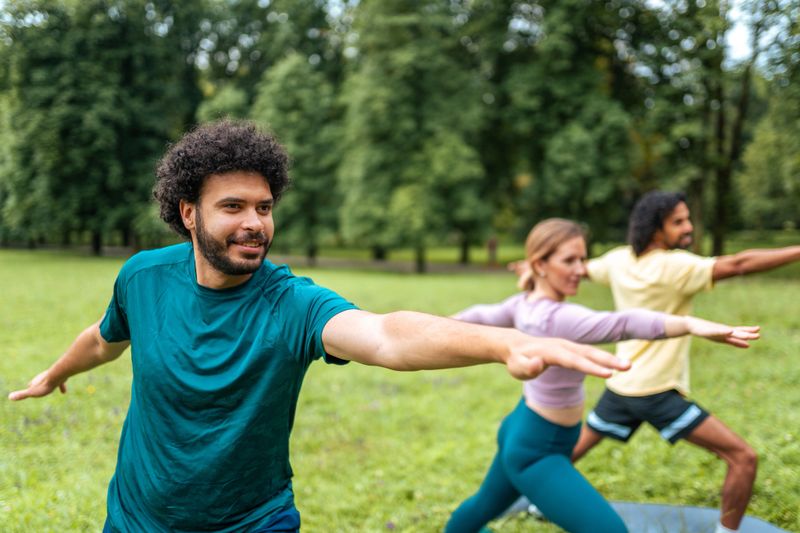 A group of smiling friends of diverse ethnicities engage in a yoga session at a lush green park, wearing casual sportswear and demonstrating a balance pose.