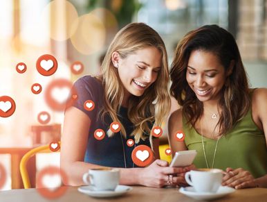 Two women happily sharing content on a phone with floating love icons around them.