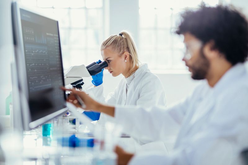 A Middle Eastern male and Caucasian female, clad in white lab coats, concentrate on analyzing specimen under a microscope, amidst scientific tools in a bright laboratory setting.