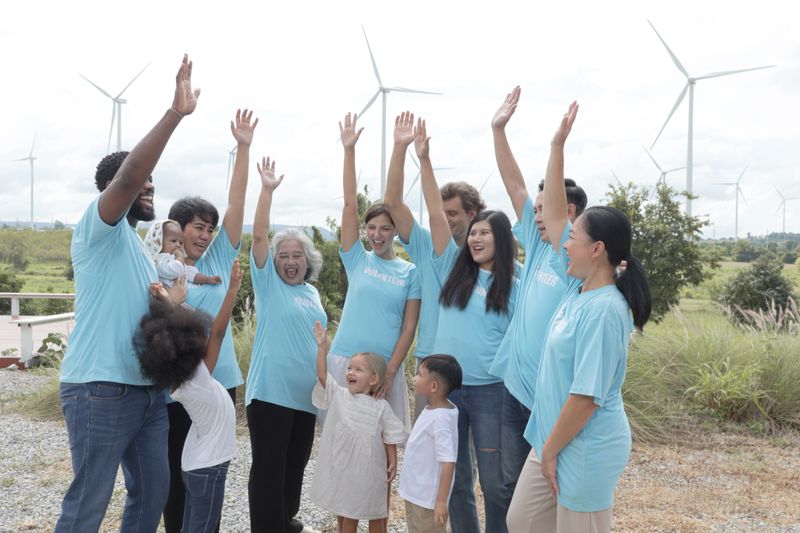 Team of diversity volunteers raising hands before collecting trash at wind turbine farm. Volunteers ready to clean up meadow with garbage collection at turbines park. Ecology and environment