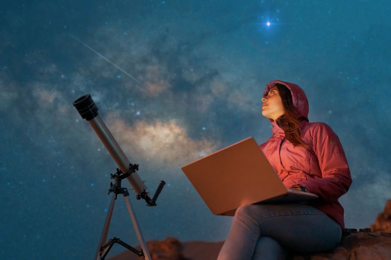 portrait woman sitting in the desert with a laptop next to a telescope at night watching the starry sky, astronomy and stargazing concept