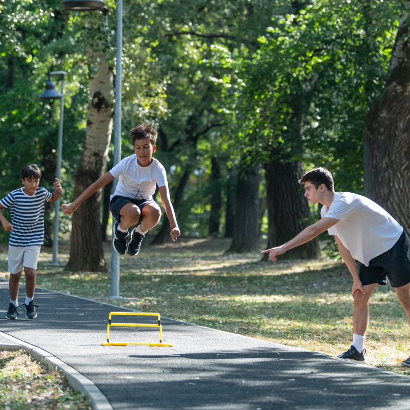Young boys jumps over hurdles outdoors during a training session with a coach. The image captures sports activities focused on improving agility and coordination in an open, natural setting.