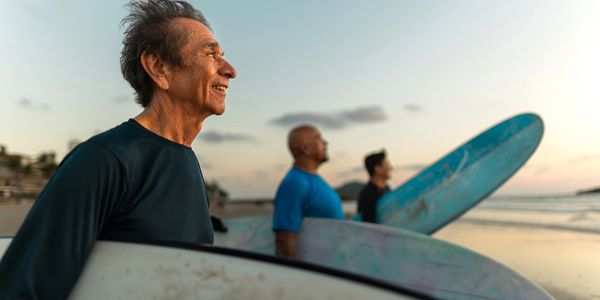 an older man on the beach holding a surfboard. enjoying his retirement