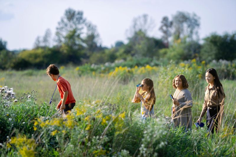 A small group of elementary students are seen exploring nature with their teacher as they take their science class outdoors.  They have magnifying glasses, binoculars and notepads as they record their findings to bring back and share with the class.