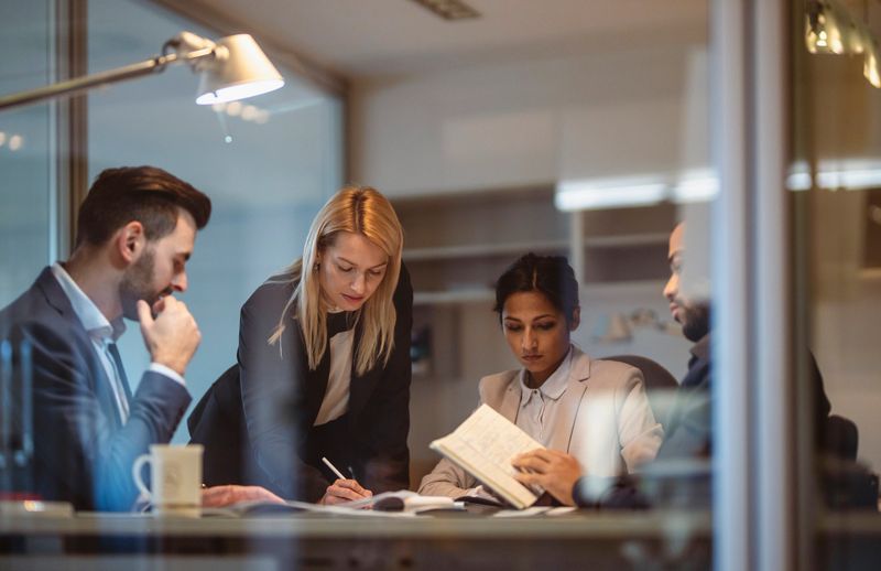 A focused group of male and female executives with diverse backgrounds collaborates on a project at a conference table in a well-lit modern office, reviewing documents and strategizing.