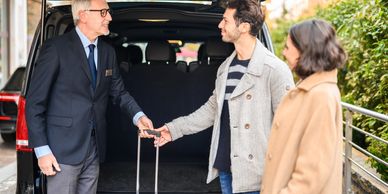 A chauffeur helps a couple with their luggage by a black vehicle.