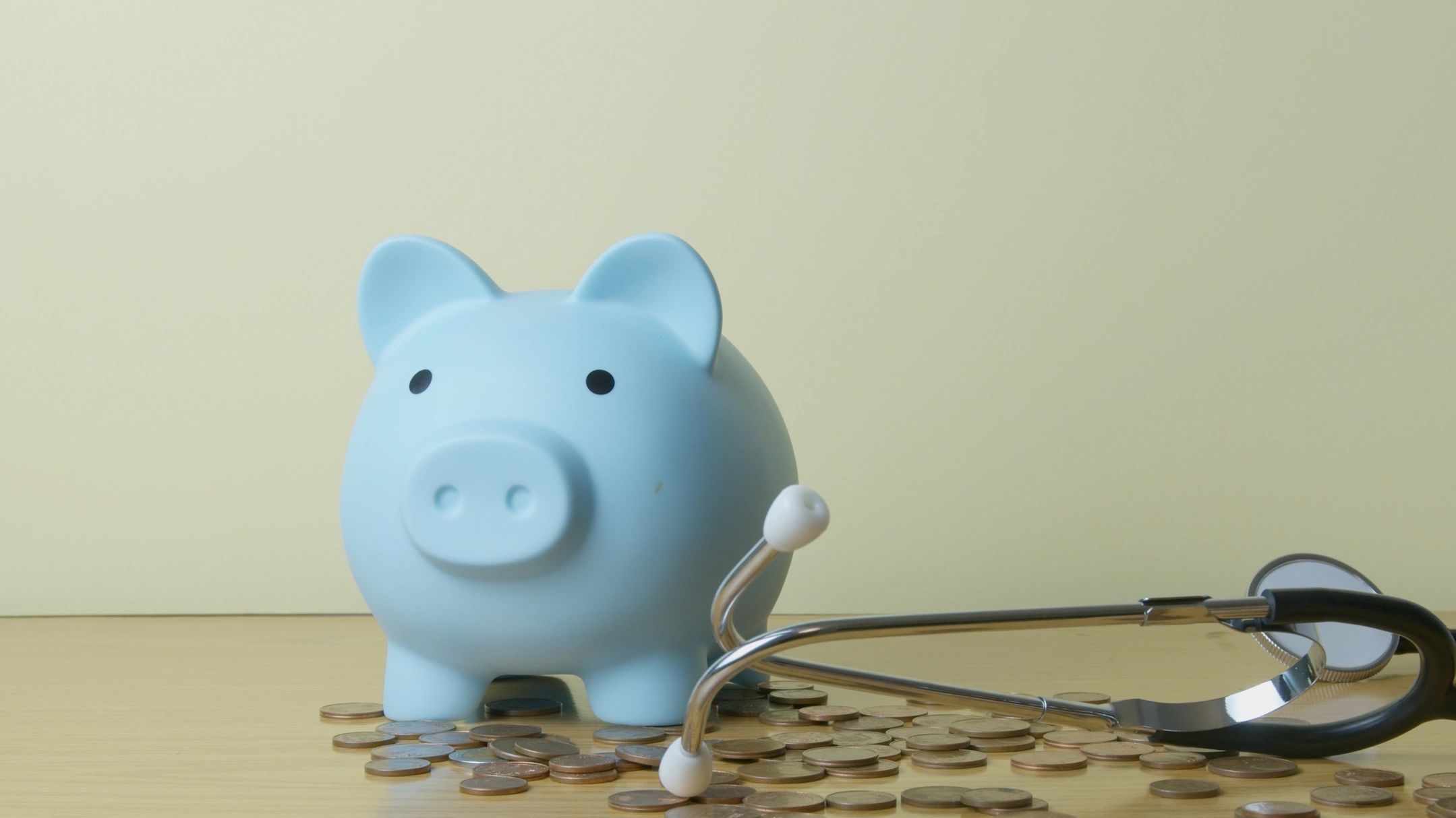 A light blue piggy bank sits on top of loose change with a stethoscope to its right. 