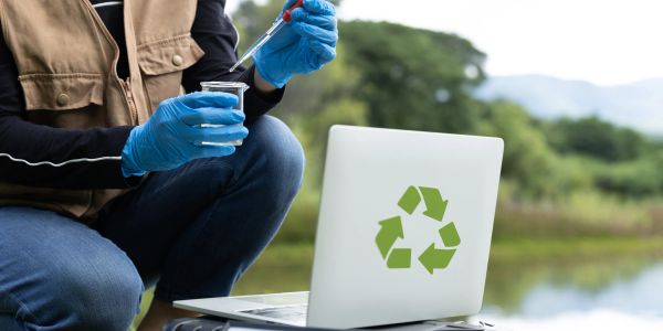 Scientist testing water outdoors with laptop showing recycling symbol.