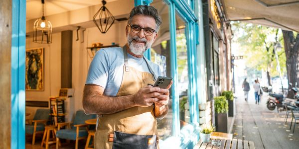 Smiling barista standing outside a coffee shop holding a smartphone.
