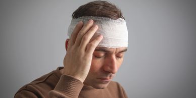 Man with a bandaged head touching his temple, eyes closed in discomfort.