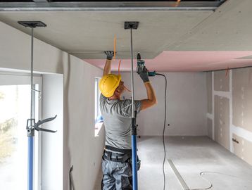 Worker installing drywall ceiling with power drill and support poles.