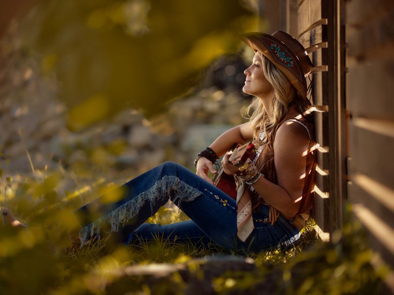 Smiling cowgirl enjoying while playing a guitar during a relaxing day by the fence on a ranch. Copy space. Photographed in medium format.