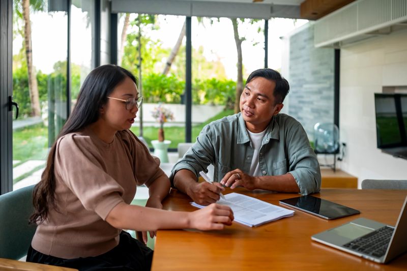 A couple sits together at a table, engaged in a serious discussion over some paperwork. The bright setting suggests a comfortable home environment, highlighting collaboration and communication.
