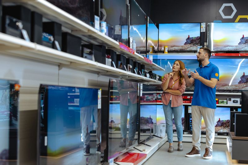 A woman consults with a male store employee in the electronics section of a retail store. They are discussing product details while standing in front of large displays.