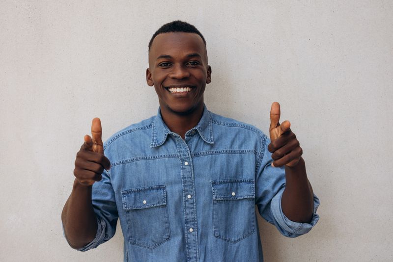 A young man in a casual denim shirt smiles brightly as he gives two thumbs up. His positive and energetic gesture adds a sense of motivation and enthusiasm. The plain background keeps the focus on his expressive facial expression and confident pose, conveying a cheerful and friendly vibe.