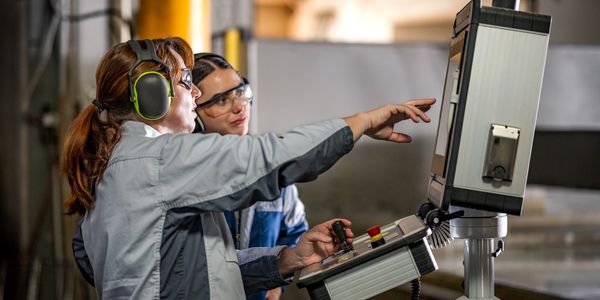 Two female workers operating a machine control panel in a factory.
