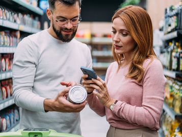 A couple shopping, checking a product and using a phone in a grocery aisle.
