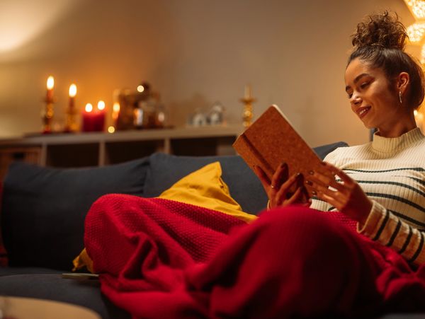 Woman reading a book wrapped in a red blanket on a cozy couch with candlelight.
