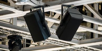 Two black speakers hanging from a metal ceiling rig in an indoor venue.